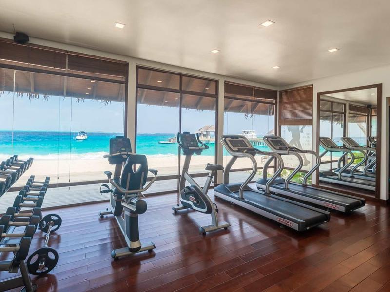 Fitnessraum mit Holzboden, Laufbändern, Hanteln und Blick auf türkisfarbenes Meer.