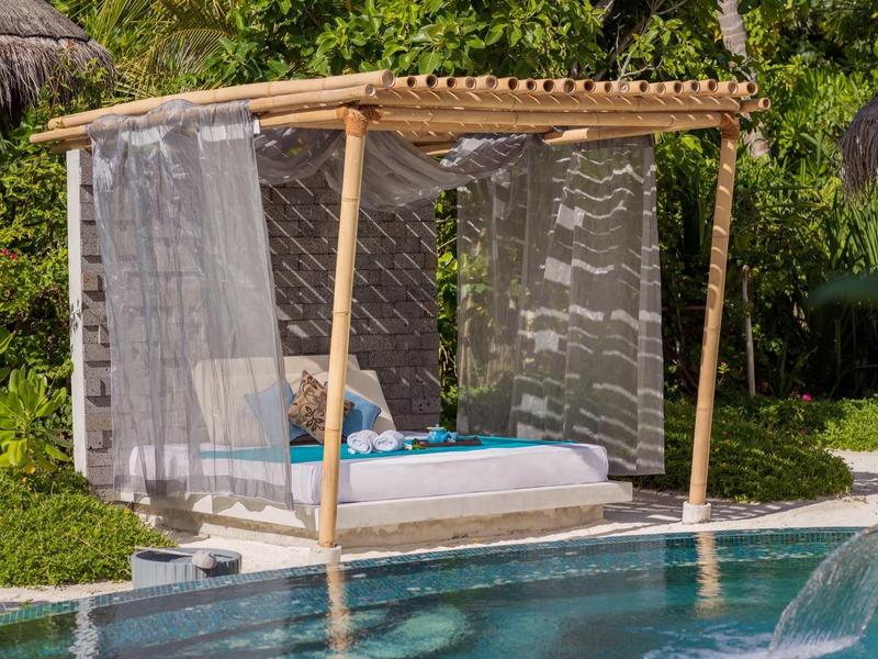 Ein gemütlicher Pavillon aus Bambus mit weißen Vorhängen und blauen Kissen neben einem Pool.