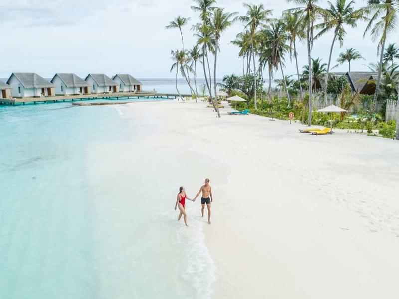 Zwei Personen laufen am weißen Sandstrand entlang, blaues Wasser links, Palmen und Wasserbungalows im Hintergrund.