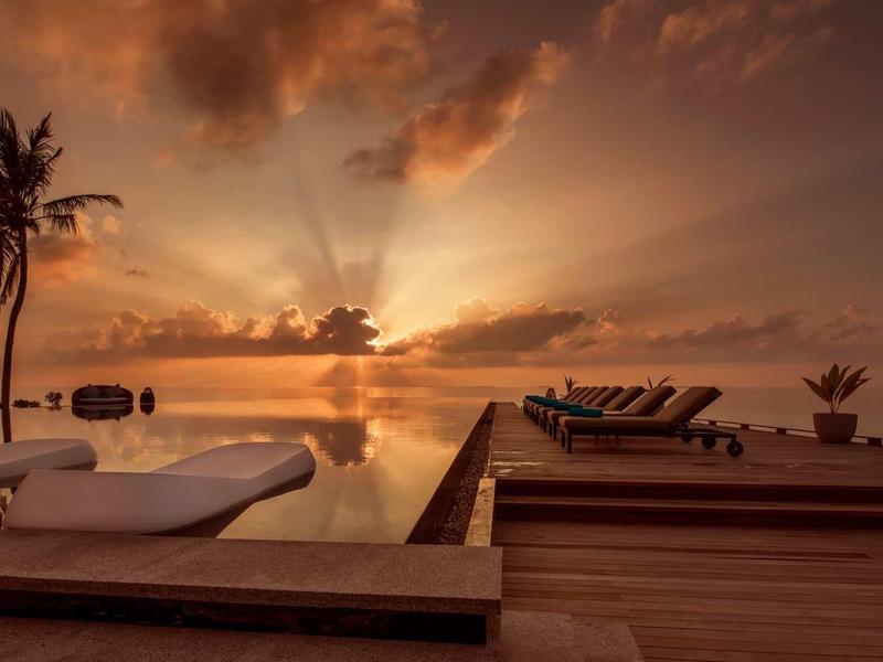 Sonnenuntergang am Strand mit Palmen, Holzdeck und Liegen im warmen Orangeton.