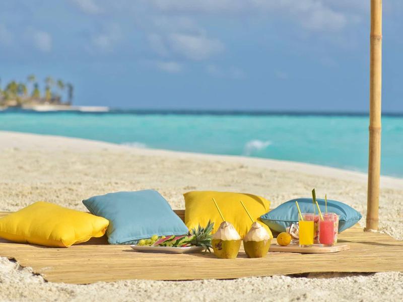 Strand mit Matratze, bunten Kissen und vier Getränken auf einem Tablett bei blauem Meer.