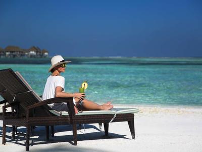 Person wearing a hat sits on a lounge chair on the beach beside turquoise sea under clear sky.