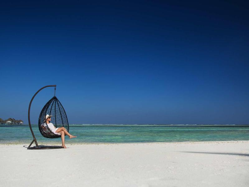Osoba siedzi w wiszącym fotelu na białej plaży z czystym niebieskim niebem i turkusowym morzem.