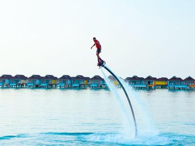 Person flies with water jetpack over calm lagoon in front of colorful water bungalows.