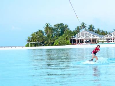Person on a flyboard over blue water near a tropical resort with palm trees.