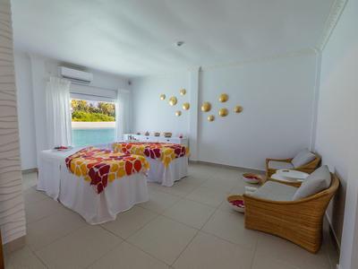 Lichte wellnessruimte met twee massagetafels en twee rotan stoelen tegen witte muren.