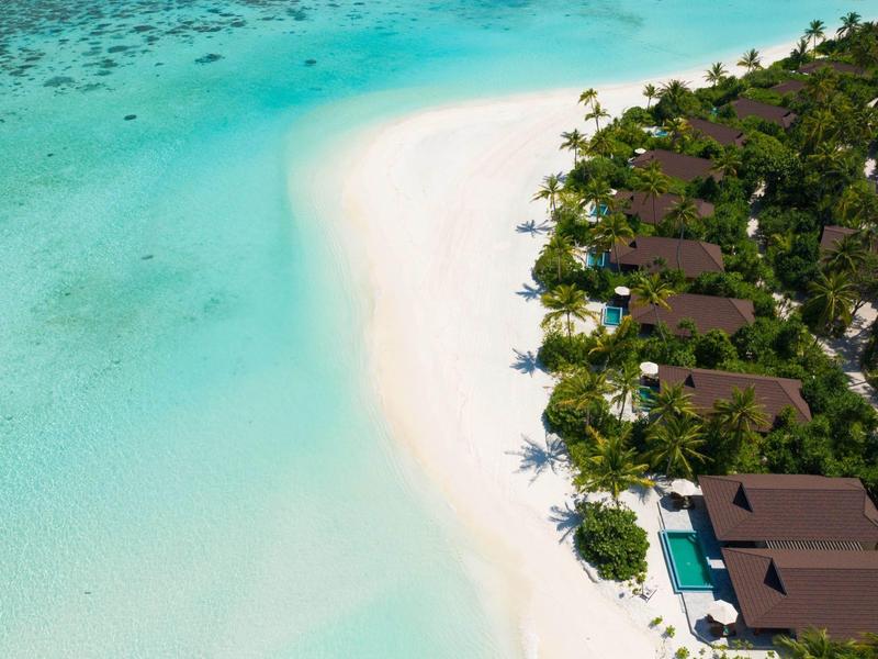 Luchtfoto van bungalows langs een wit zandstrand met turquoise water en palmbomen.
