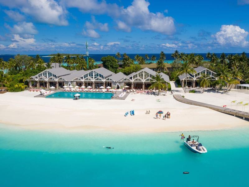 Resort met strand, zwembad en boot op turquoise zee onder blauwe lucht.