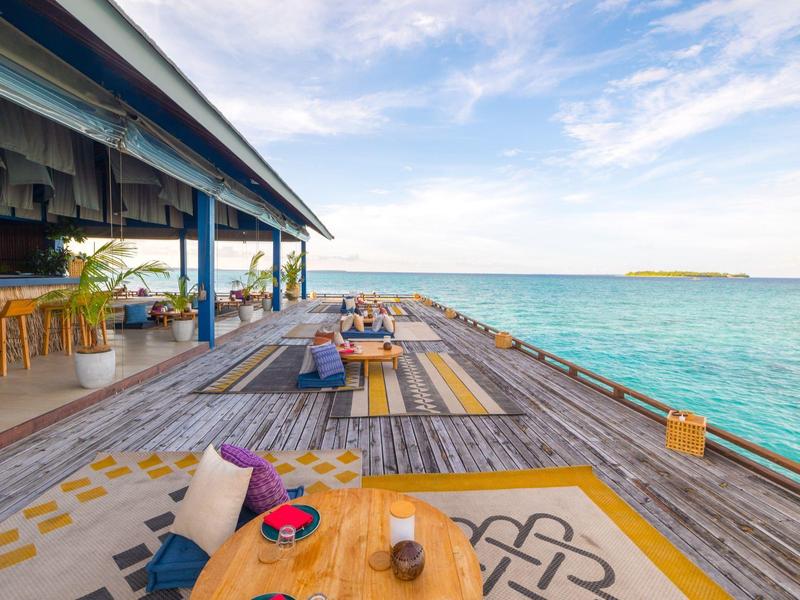 Een houten terras aan zee met kussens, tafels en een overdekt gedeelte met stoelen.