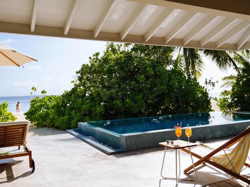 Uitzicht op een klein zwembad en ligstoelen onder een overdekt gebied met de zee op de achtergrond.
