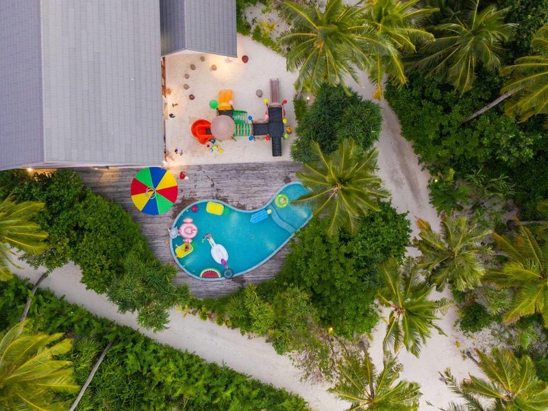 Luchtfoto van een tropisch zwembad met kleurrijke parasols omgeven door palmbomen.