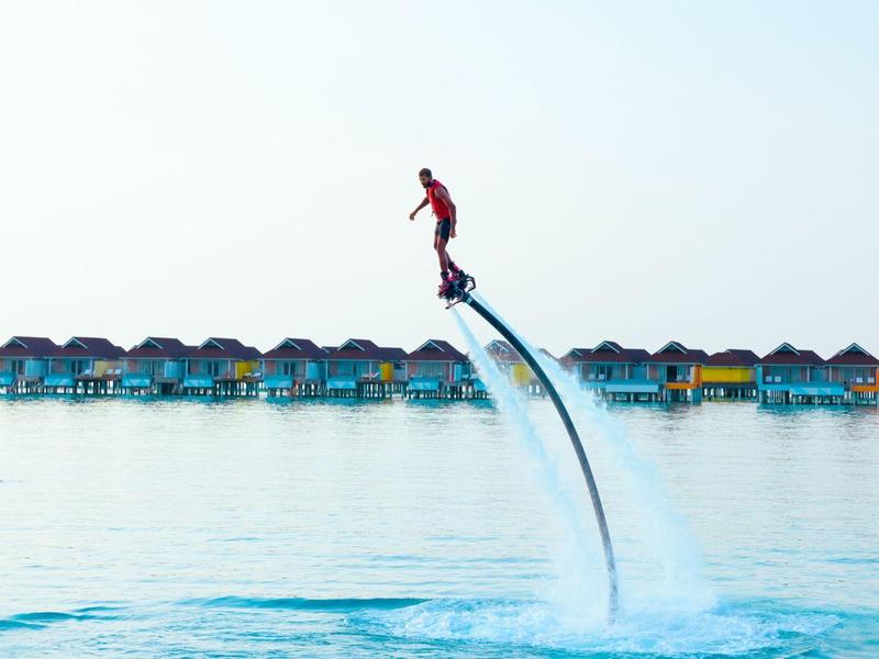 Persoon vliegt met waterjets voor kleurrijke waterbungalows boven kalme zee.