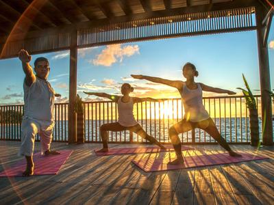 Trzy osoby ćwiczą jogę na tarasie o zachodzie słońca z widokiem na wodę.