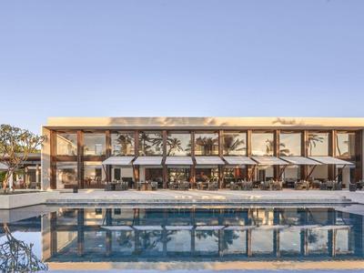 Bâtiment d'hôtel moderne avec salon ouvert et grande piscine au premier plan sous ciel dégagé.