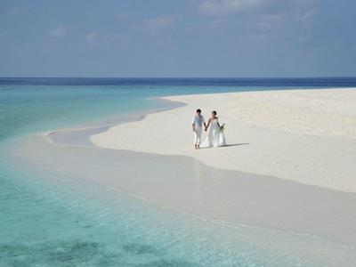 Couple en vêtements blancs marchant sur une plage de sable calme en bordure d'eau turquoise claire.