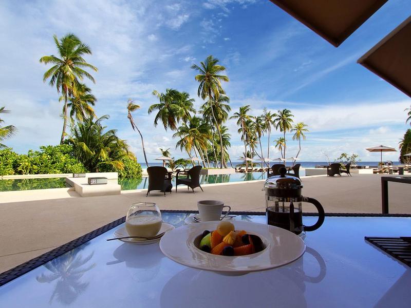 Table avec café et fruits sur une terrasse avec palmiers et vue sur la mer.
