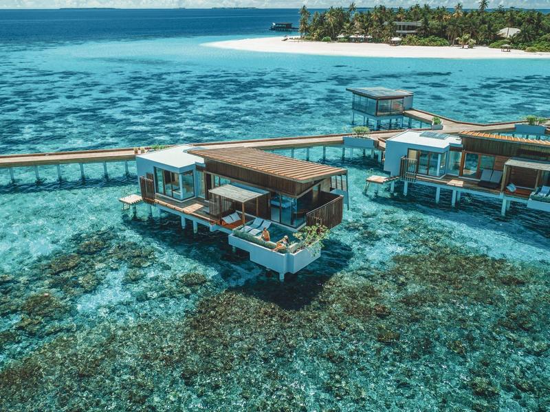 Bungalows sur pilotis avec passerelles en bois sur une eau turquoise claire près d'une île aux palmiers.
