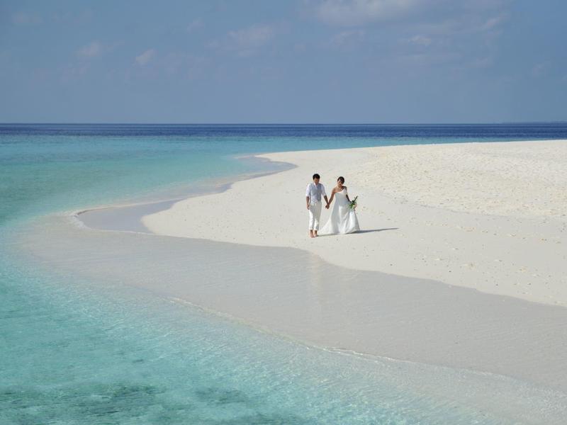 Couple en vêtements blancs marchant sur une plage de sable calme en bordure d'eau turquoise claire.