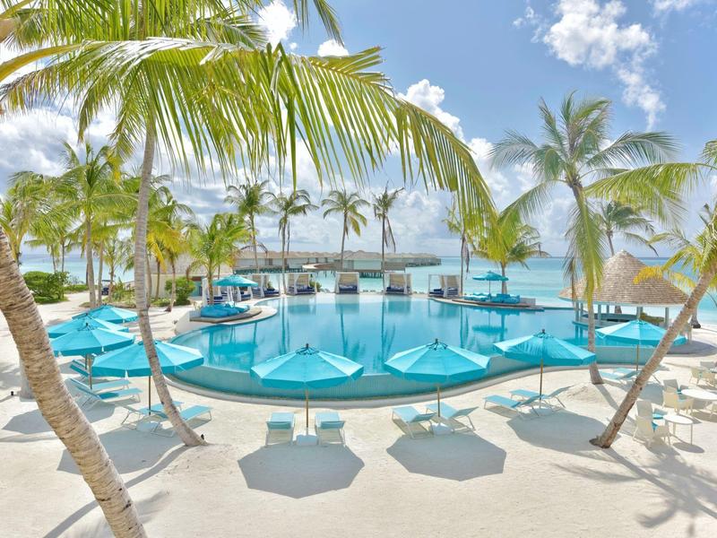 Strand mit weißem Sand, Palmen und blauen Sonnenschirmen vor türkisfarbenem Meer und klarem Himmel.