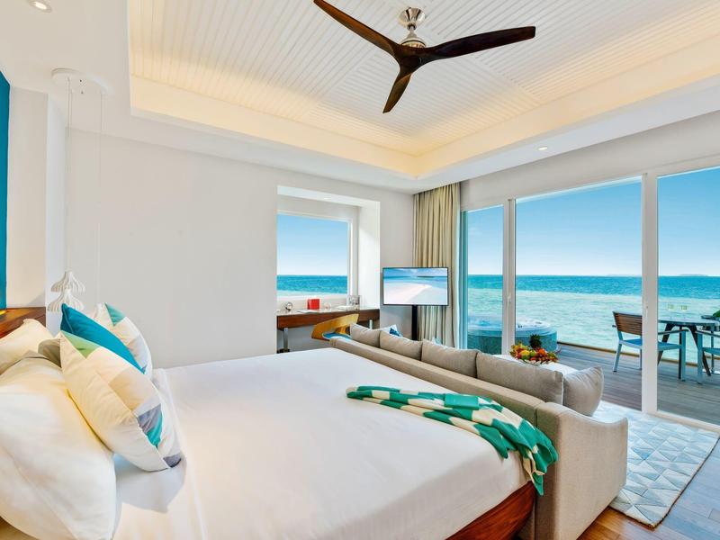 Modernes Schlafzimmer mit großem Bett, Deckenventilator und Blick auf Meer durch große Fenster.