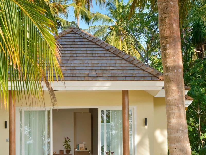 Kleines Haus mit Spitzdach und Glastüren, umgeben von Palmen unter blauem Himmel.