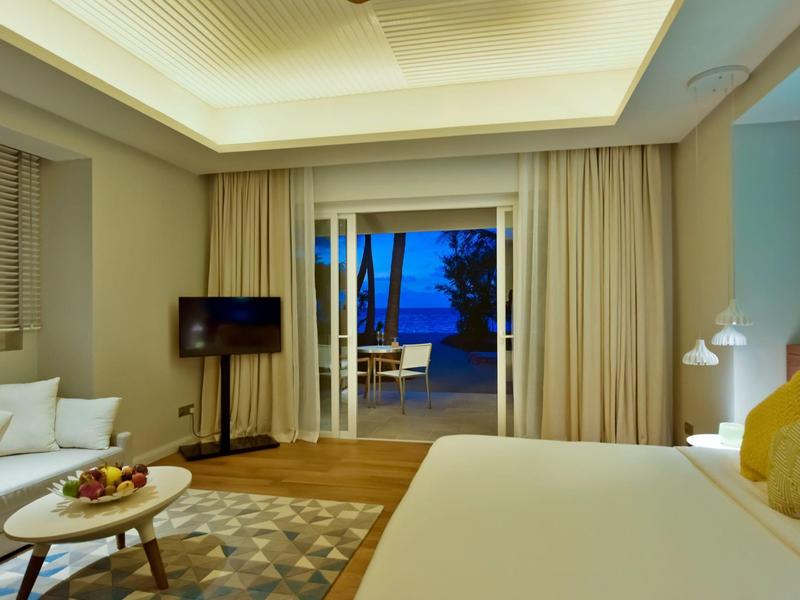 Modernes Schlafzimmer mit großem Bett, Sofa, Teppich, Fernseher und offener Terrassentür zum Abendhimmel.