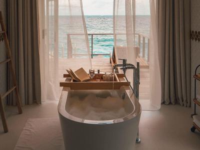 Badewanne mit Schaum vor Balkon mit Meerblick durch transparente Vorhänge.