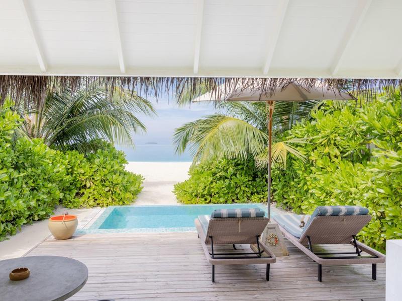 Blick von einer Terrasse mit Pool auf Strand und Meer, umgeben von Palmen und Grün.