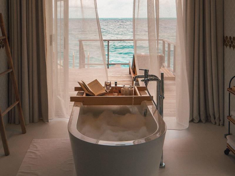 Badewanne mit Schaum vor Balkon mit Meerblick durch transparente Vorhänge.