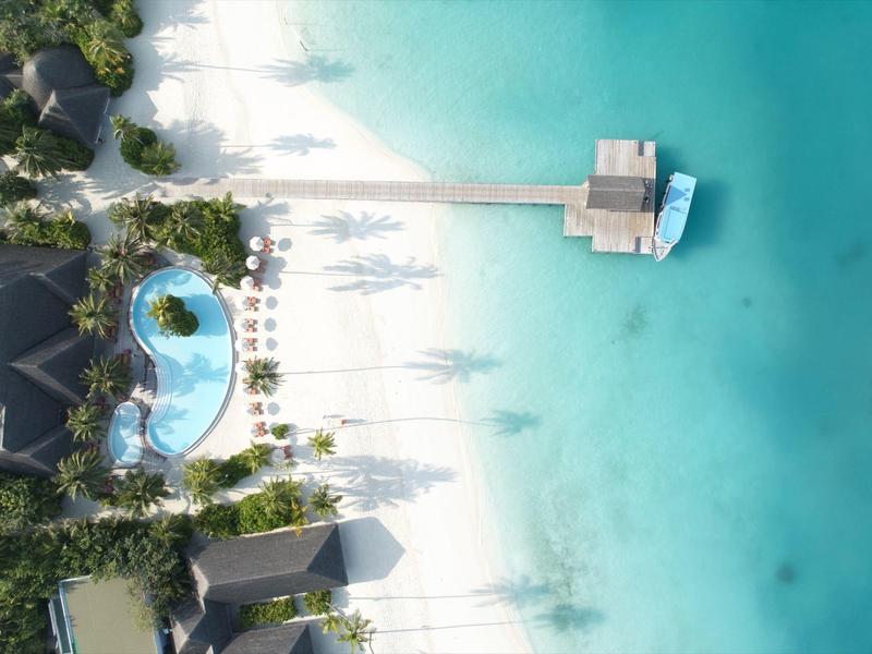 Vogelperspektive auf einen Resort-Pool mit Sonnenschirmen und einem Steg über türkisfarbenes Wasser.