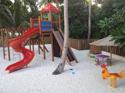 Kinderspielplatz mit roter Rutsche, Sandboden und bunten Sitzgelegenheiten neben Palmen.