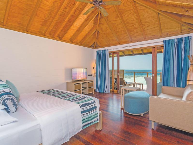 Helles Hotelzimmer mit Holzboden, Bett, Sofa und Balkon mit Meerblick.