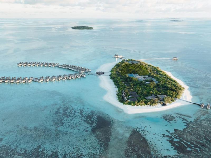 Una isla tropical con vegetación exuberante y bungalows sobre el agua, rodeada de aguas cristalinas y turquesas.