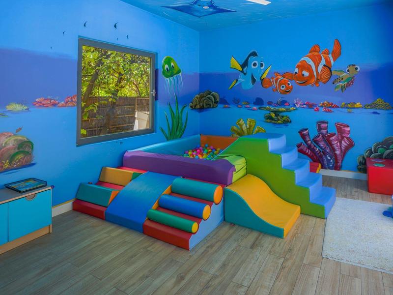 Habitación de juegos infantil colorida con tobogán y murales marinos, ventana luminosa, suelo de madera.