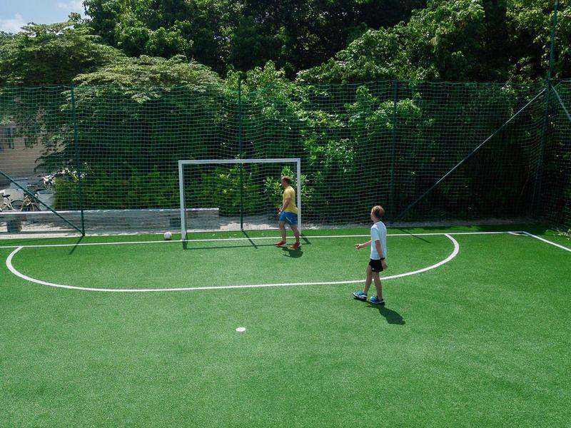 Dos niños juegan en un pequeño campo de fútbol con césped artificial verde y porterías al aire libre.