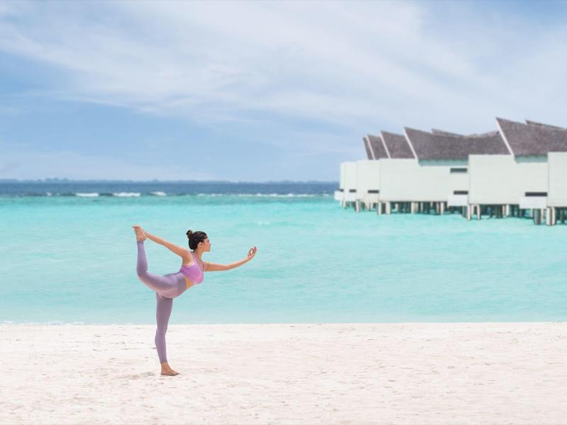 Mujer haciendo yoga en playa de arena blanca con agua turquesa y bungalows al fondo.
