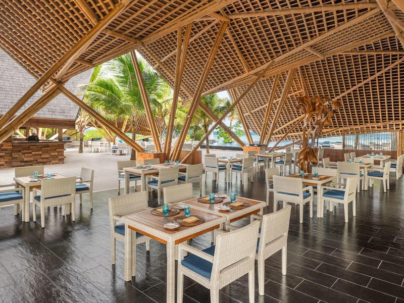 Restaurante abierto con techo de madera, mesas y sillas blancas en entorno tropical.