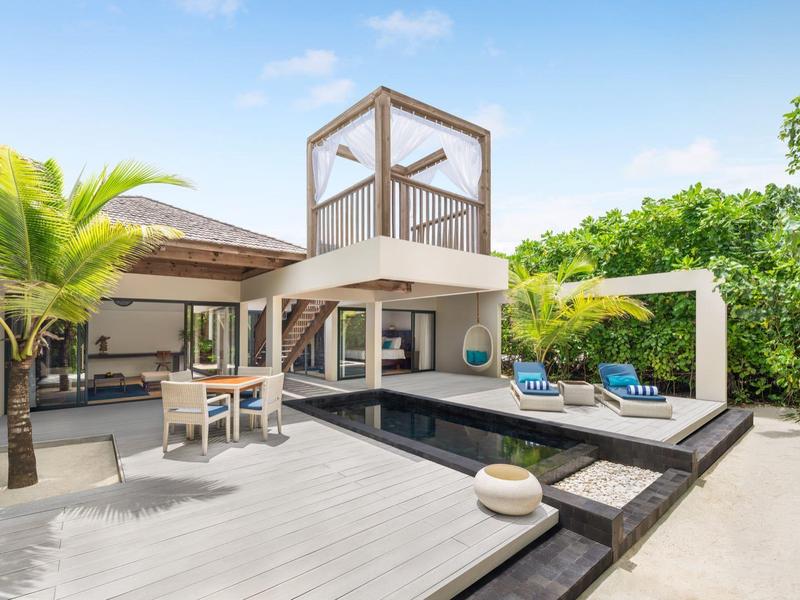 Área de piscina moderna en hotel con tumbonas, mesa de comedor y vegetación tropical bajo sol.