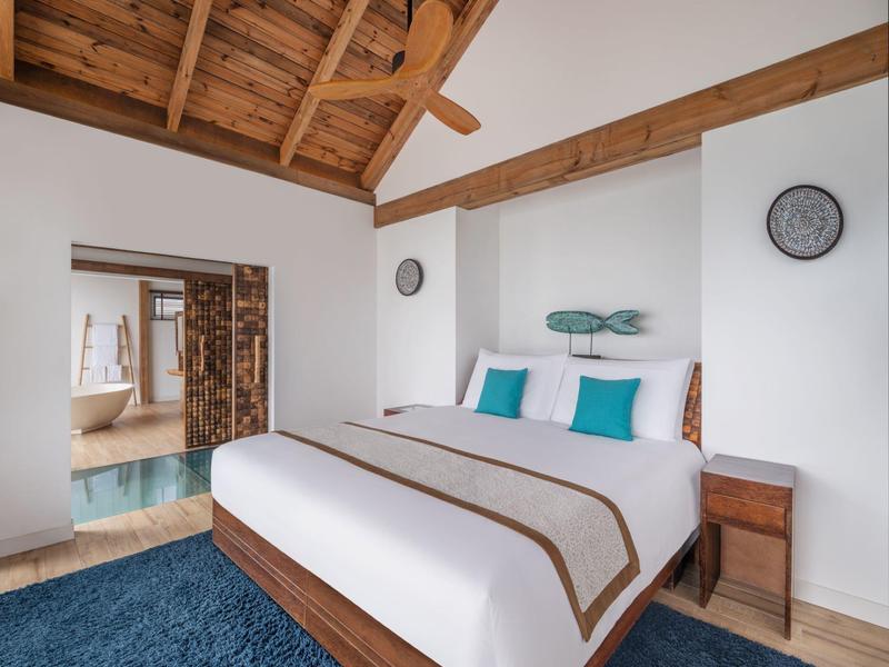 Dormitorio con cama doble, techo de madera inclinado y decoración moderna en tonos neutros y azules.