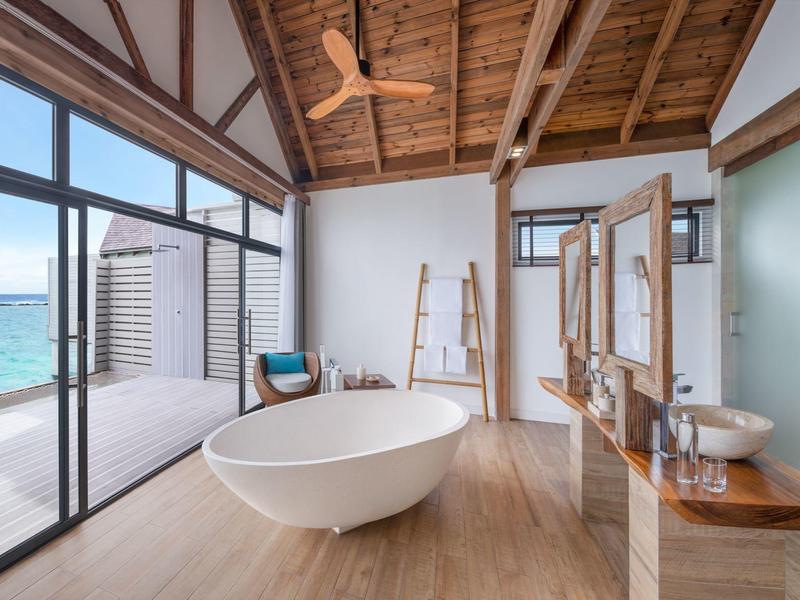 Baño luminoso con bañera independiente, suelo de madera y vista al mar a través de grandes ventanas.
