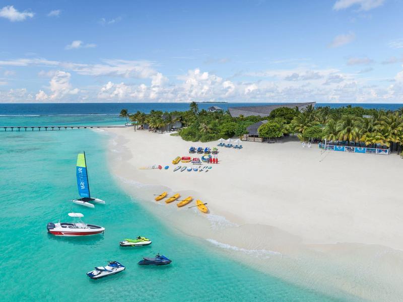 Playa tropical con arena blanca, laguna azul, equipos de deportes acuáticos y fondo de isla verde.