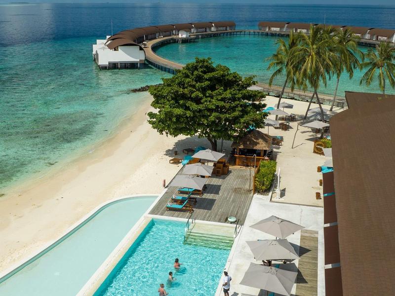 Ein tropischer Strand mit klarem Wasser, Pools, Palmen und einer Holzplattform im Meer.