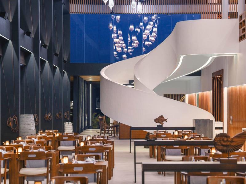 Modernes Restaurant mit Holzstühlen, Tischen und einer großen weißen Spiraltreppe im Hintergrund.