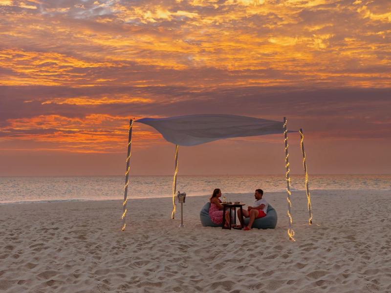 Zwei Personen sitzen unter einem Baldachin am Strand bei Sonnenuntergang mit buntem Himmel.