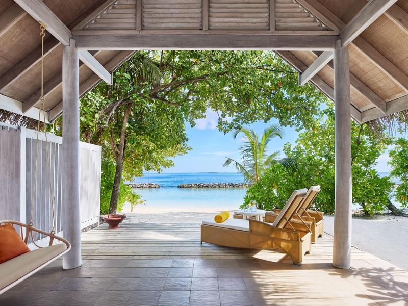 Blick von einer überdachten Terrasse auf einen tropischen Strand mit Liegestuhl und türkisfarbenem Meer.