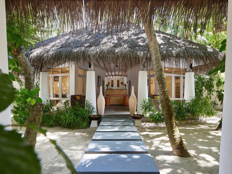 Ein Weg führt zu einer tropischen Hütte mit Strohdach, umgeben von Sand und Palmen.