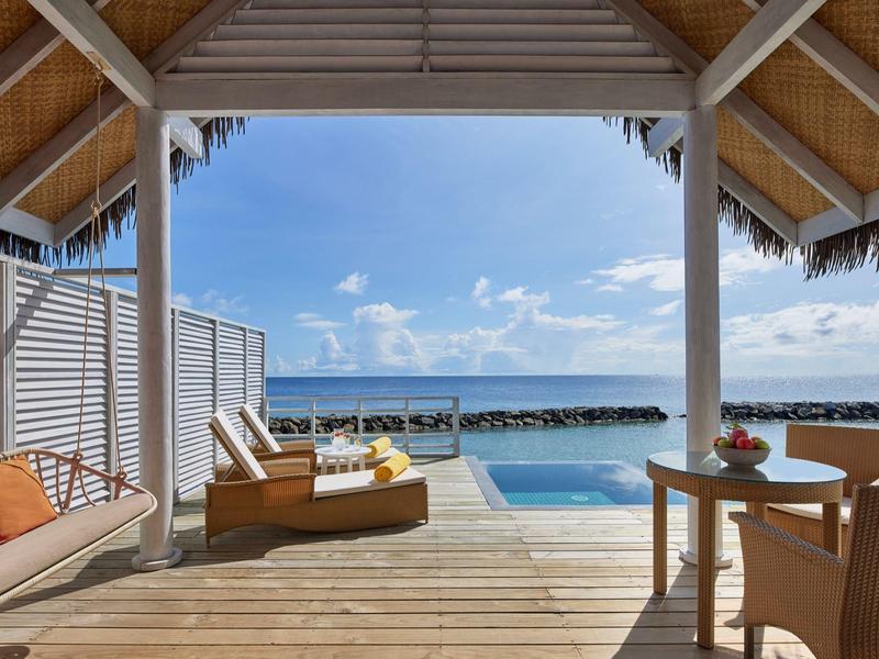 Blick von einer überdachten Terrasse mit Liegestühlen und Tisch auf das Meer und den Pool.
