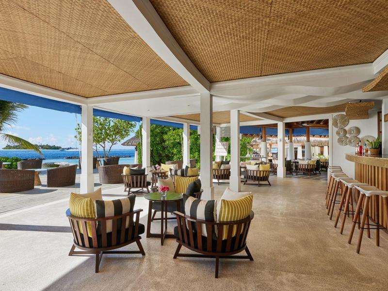 Offener Hotel-Loungebereich mit Stühlen, Tischen und Bar, direkter Blick auf den Strand und Palmen.