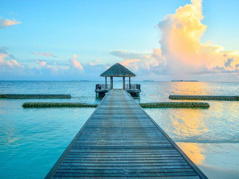 Ein Steg führt zu einer kleinen über dem Wasser thronenden Hütte bei Sonnenuntergang.