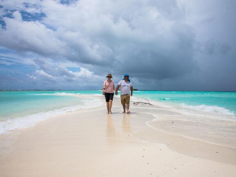 Zwei Personen gehen am weißen Sandstrand entlang in Richtung türkisfarbenes Meer unter bewölktem Himmel.